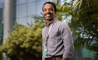 Homem sorrindo, com roupa social e crachá no pescoço, em frente a um prédio com fachada de vidro.