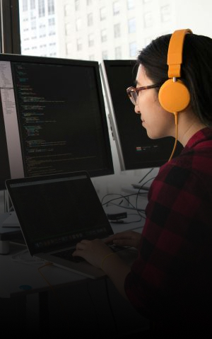 Mulher programando, ela está de frente para uma janela e três telas.