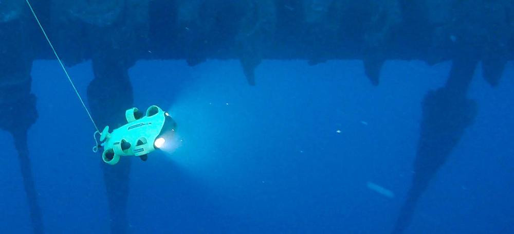 Fotografia subaquática de um minidrone usado pela Petrobras. Ele está preso a um fio e tem luzes frontais ligadas.