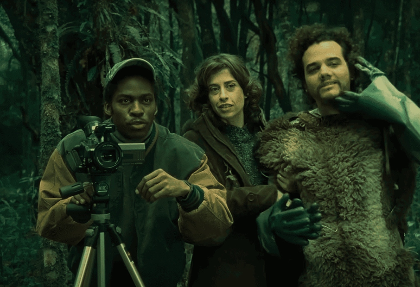 Fernanda Torres, Wagner Moura (caracterizado com pele de animal) e Lázaro Ramos (operando uma câmera de vídeo) em uma floresta, em cena do filme "Saneamento Básico, O Filme".
