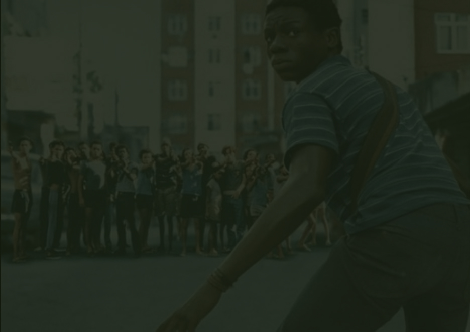 Buscapé olha para trás com receio enquanto um grupo armado o observa ao fundo, em cena de