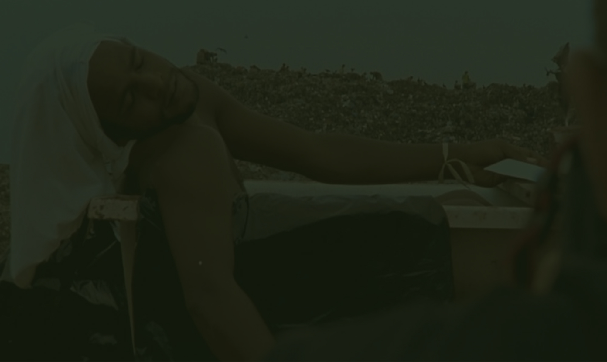 Um homem com a cabeça coberta por um pano branco repousa em uma banheira em meio a um lixão, em