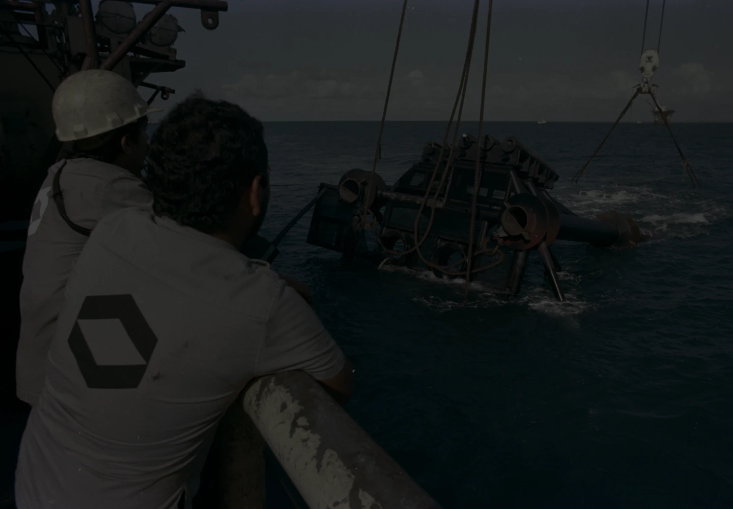 Photo of a metal structure being lifted from the sea by a Petrobras platform. On the platform, there are two employees.