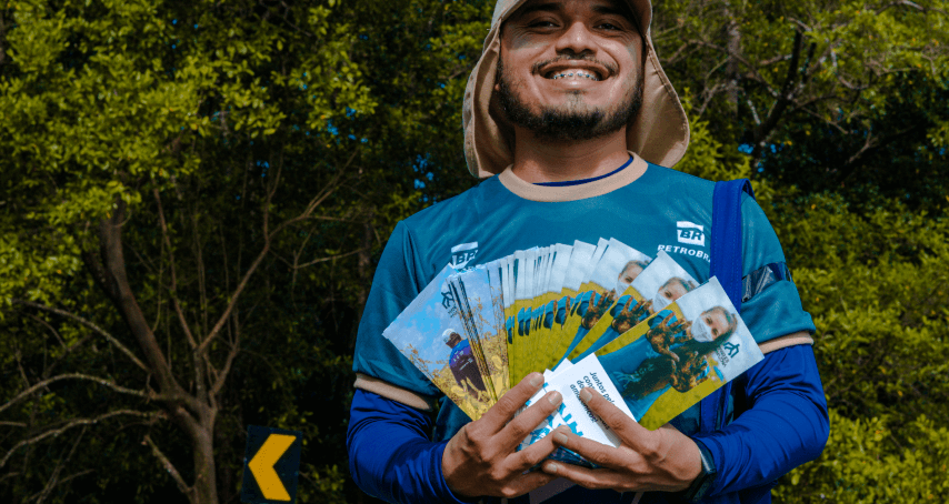 Photo of a man smiling in front of a forest, holding several flyers for the “Mangues da Amazônia” project and wearing a Petrobras T-shirt.