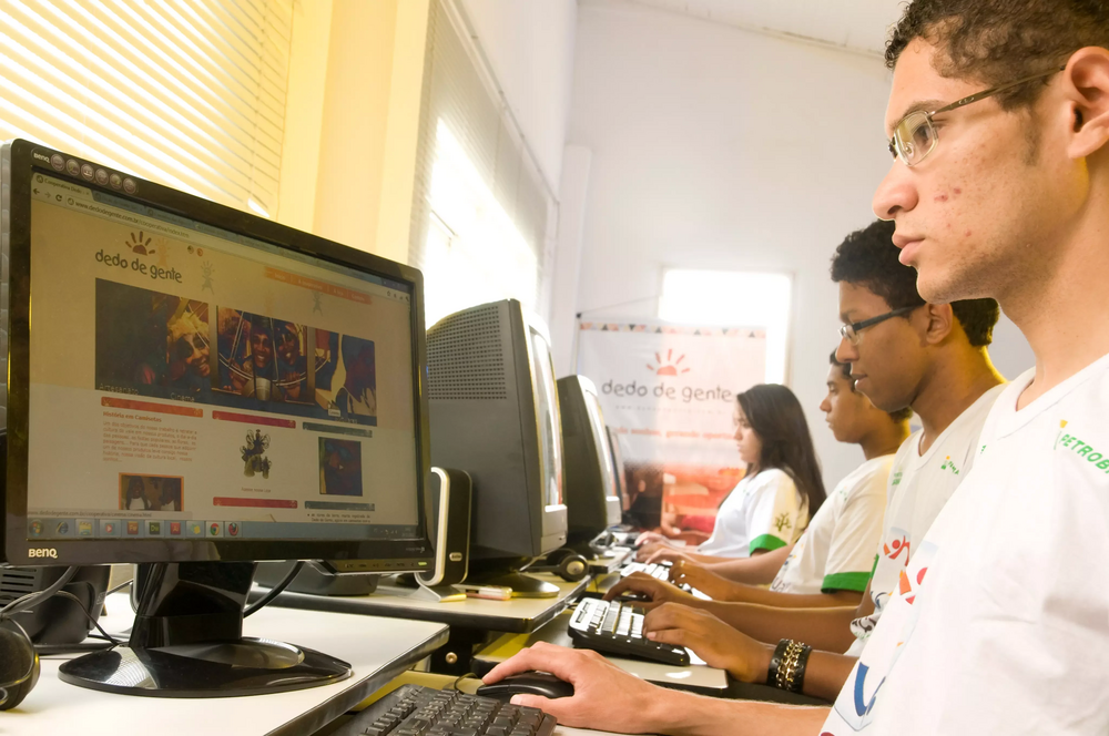 Quatro jovens sentados em frente a computadores, em uma sala de informática.