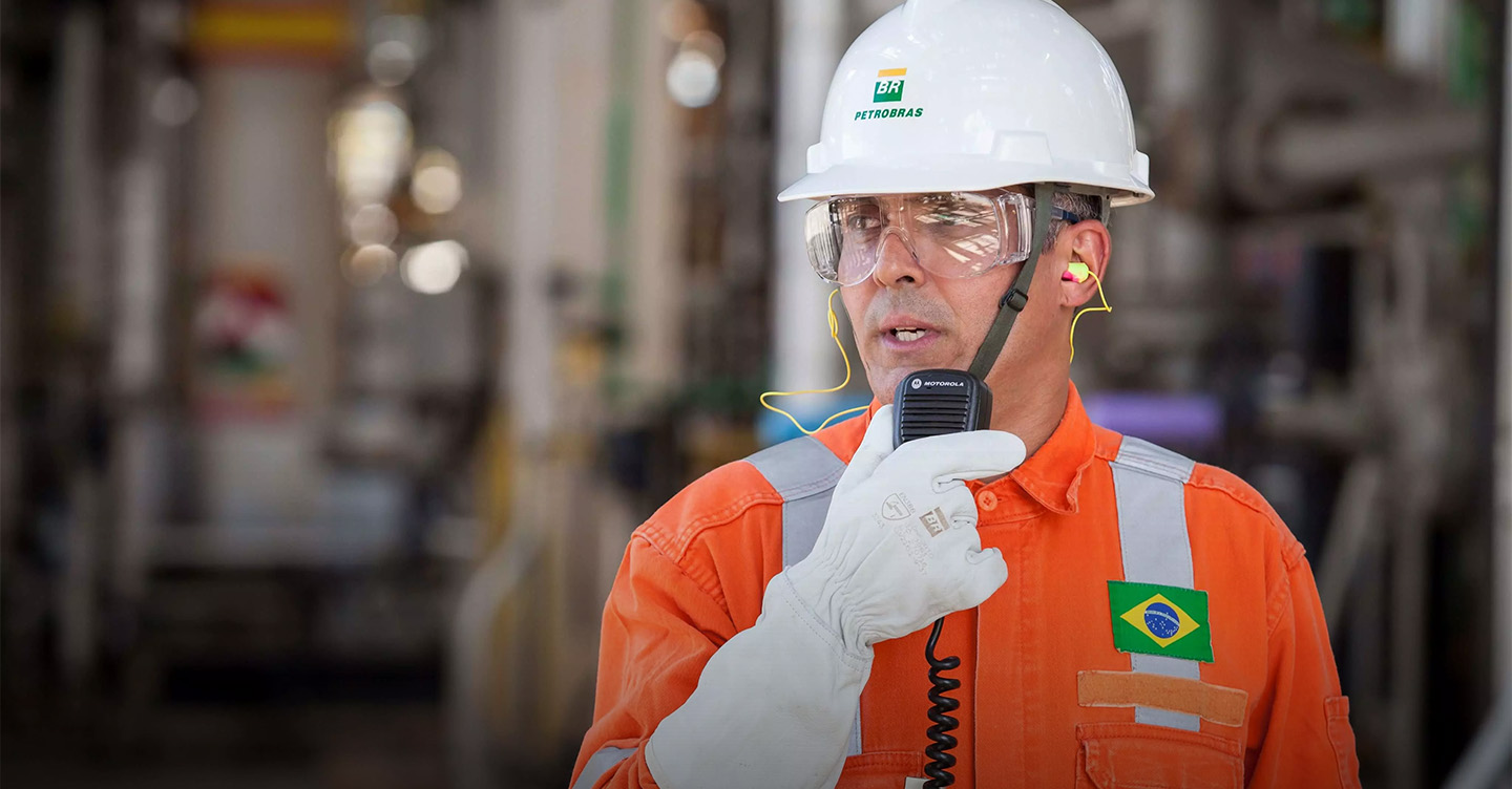 Petrobras professional wearing a white helmet and orange overalls, speaking into a radio transmitter.