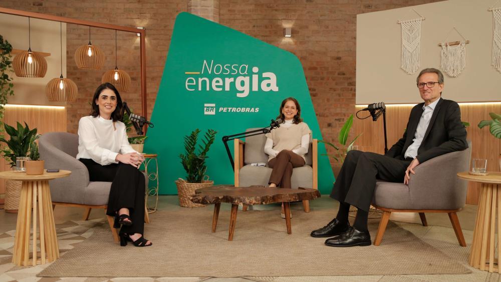 Duas mulheres e um homem sorriem para a câmera no cenário do podcast Nossa Energia, da Petrobras.