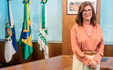 Magda Chambriard, nova presidente da Petrobras, sorri para a foto. Atrás dela estão as bandeiras do Rio de Janeiro, do Brasil e da Petrobras.