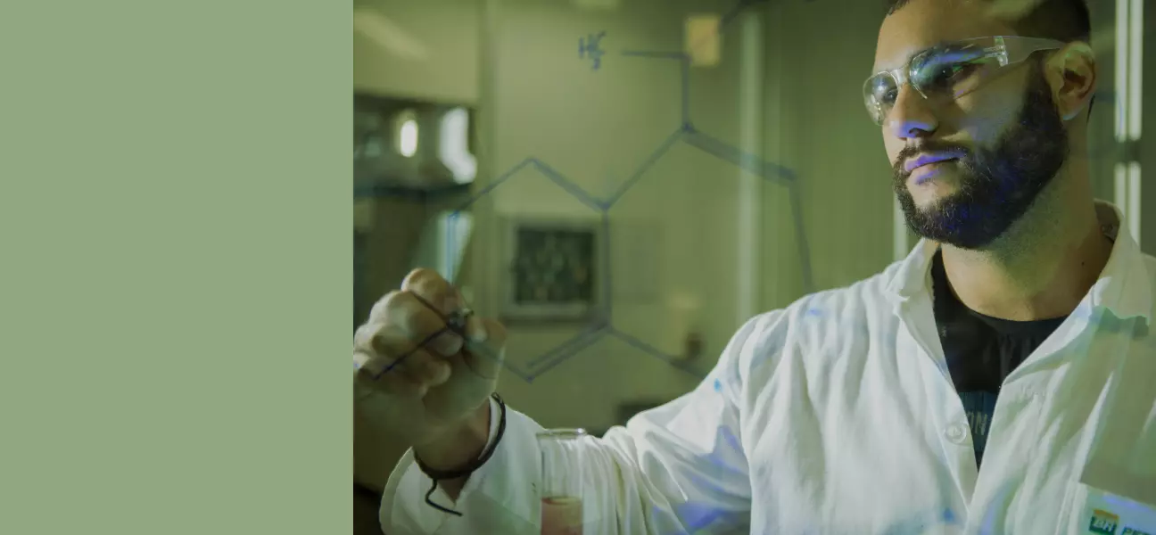 Cientista da Petrobras, com jaleco branco e óculos de proteção, escrevendo uma fórmula química em um vidro transparente.