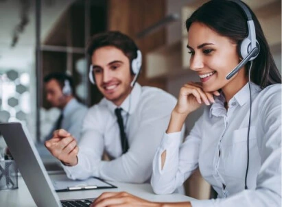 Fotografia de um homem e uma mulher, ambos usando camisa social, sentados em frente a um computador com fone de ouvido com microfone.